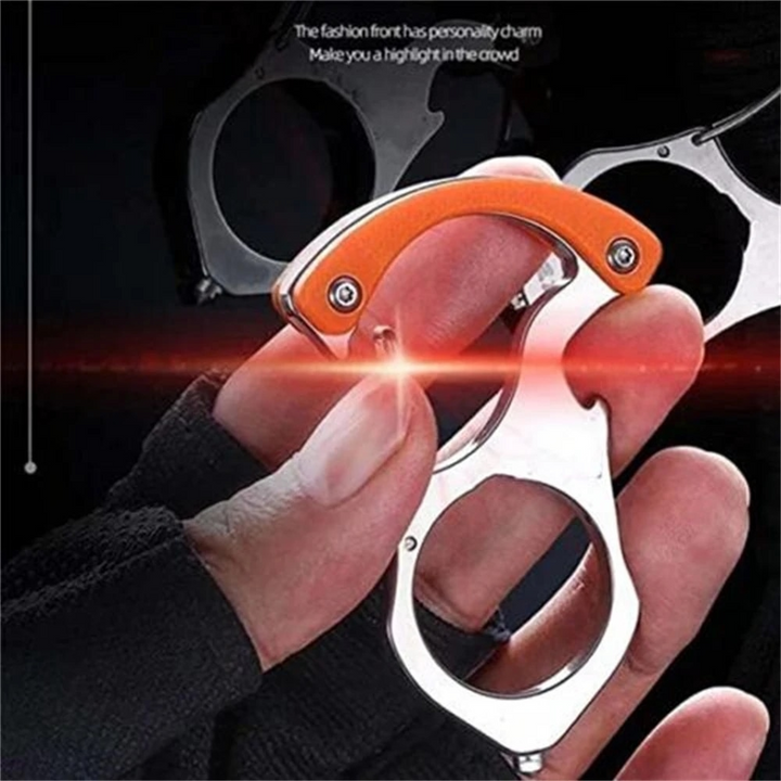 Hånd holder en blank stålnøglering med orange greb tæt på kameraet, mens flere lignende modeller svagt anes i mørk baggrund. 1