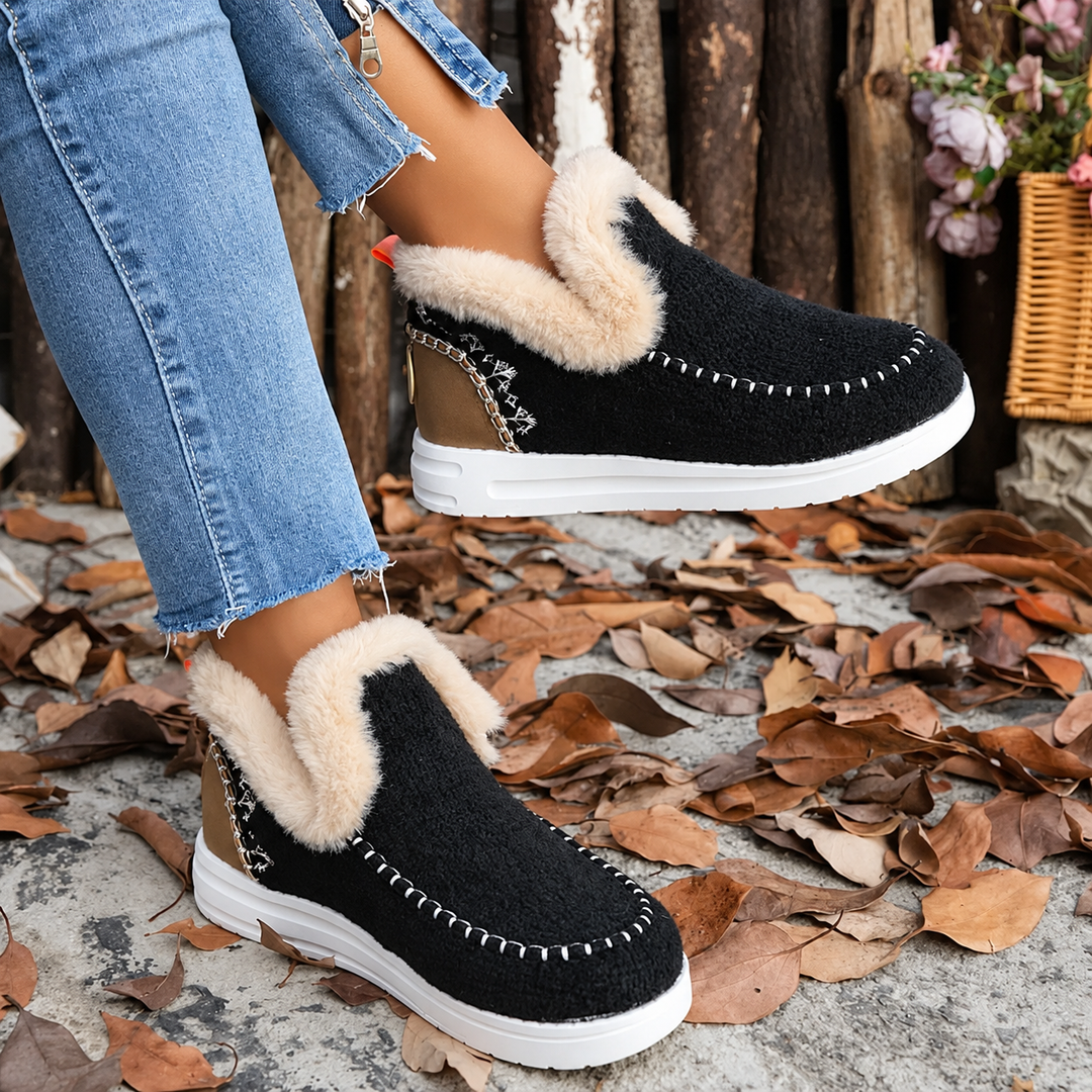 Sorte uldne slip-on sneakers med cremefarvet plysskaft og hvide sting, båret med jeans blandt efterårsblade. 1