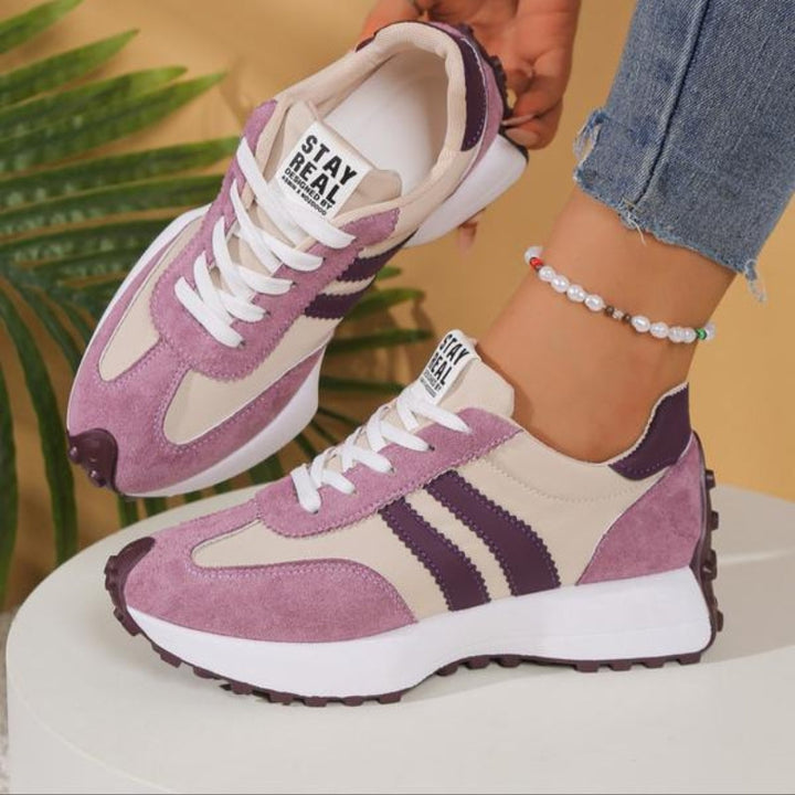 Lilla og beige sneakers med mørklilla striber og hvide snørebånd, hvor den ene sko løftes af en model 11