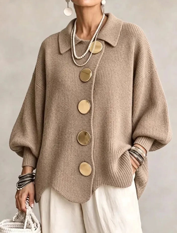 Model i sandfarvet ribstrikket cardigan med spids krave og store guldfarvede knapper, set forfra med hvide bukser. 7