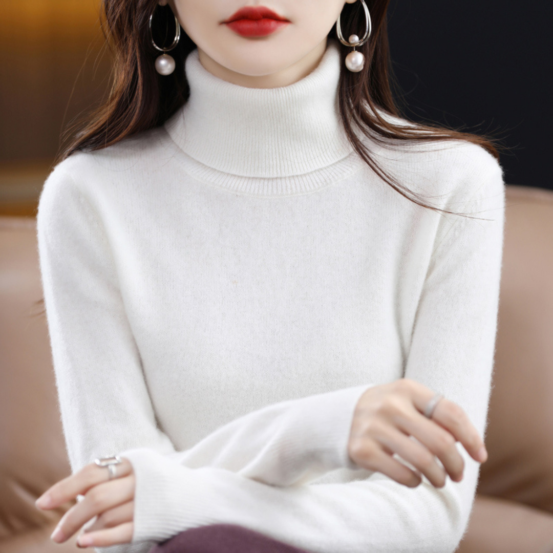 Model i hvid rullekravesweater med høj krave og hænderne krydset foran kroppen, siddende i en sofa. 12