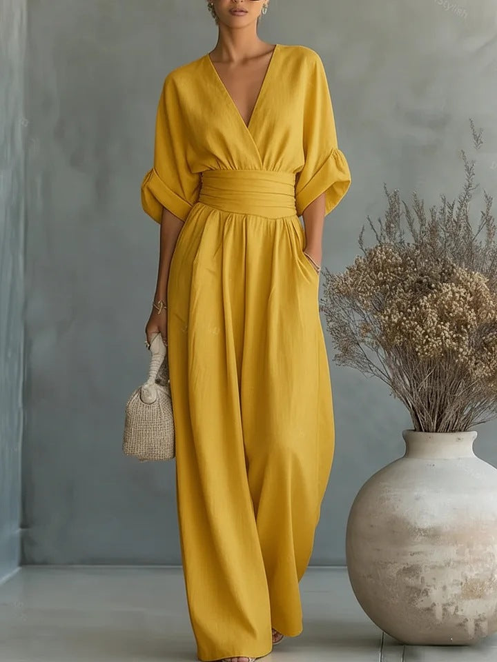 Jumpsuit i gul set forfra på model med V-udskæring, foldede ærmer og vide ben foran grå væg 3