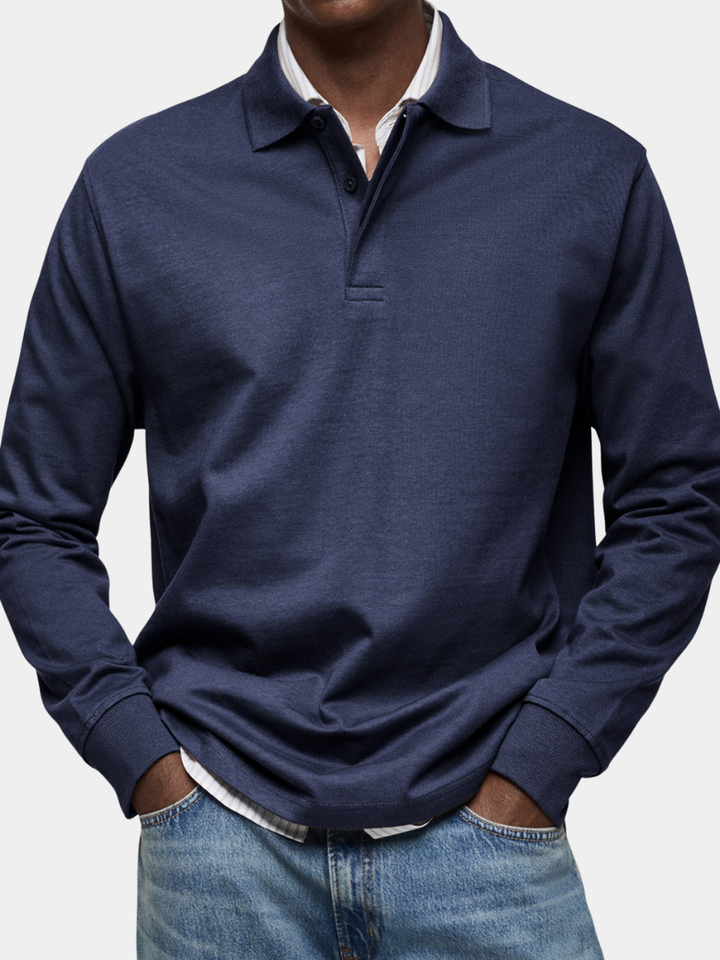 Mørkeblå langærmet poloshirt med ribkant ved ærmerne, båret af en model over en stribet skjorte og med hænderne i jeanslommerne. 1