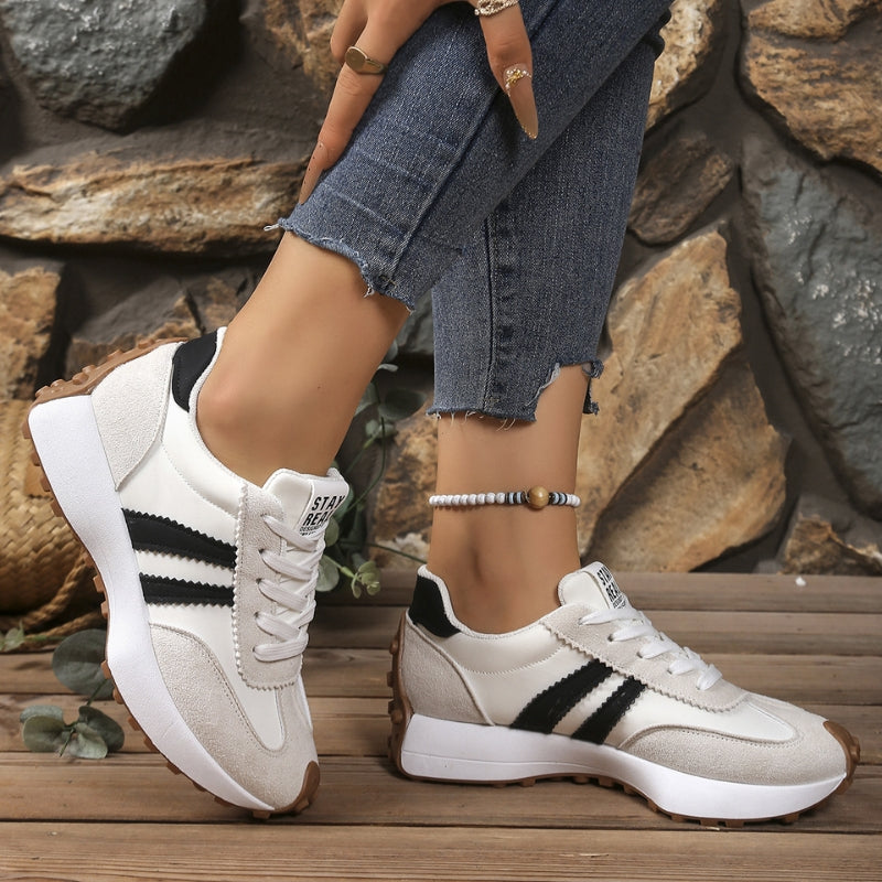 Hvide sneakers med beige ruskindsdetaljer og sorte striber båret af en model foran stenvæg 5