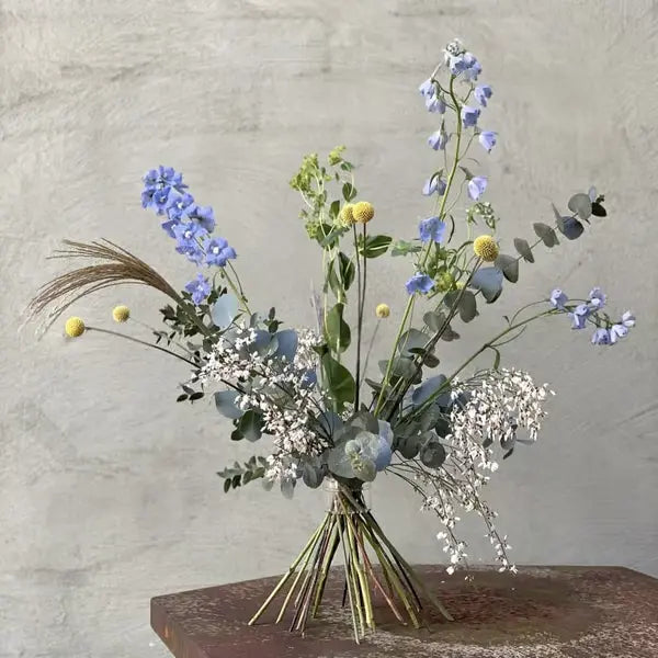 Frit stående buket med blå rittersporer, gule trommestikblomster, eukalyptus og hvide smådele foran en grå betonvæg. 12