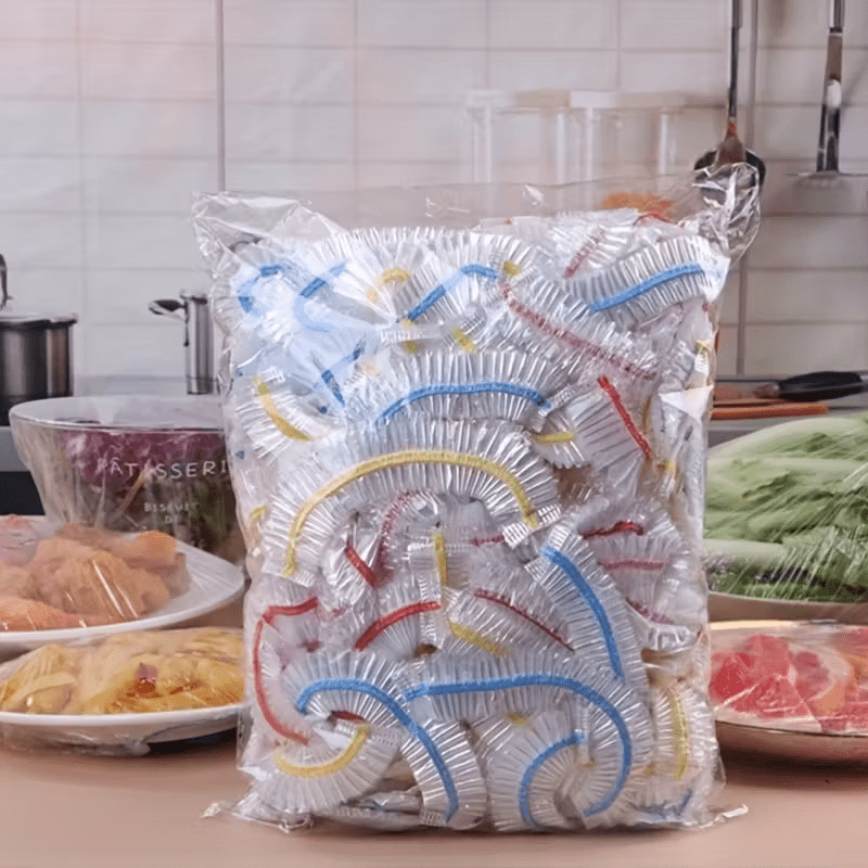 Stor gennemsigtig pose fyldt med elastiske dækkapper med blå, gule og røde kanter, stillet på et køkkenbord foran madfade. 1