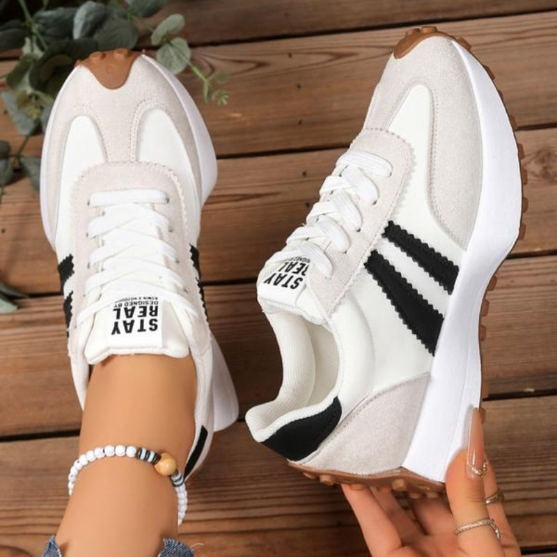 Hvide sneakers med sorte striber og beige ruskindsdetaljer, set ovenfra på trægulv med en hånd, der holder den ene sko 10
