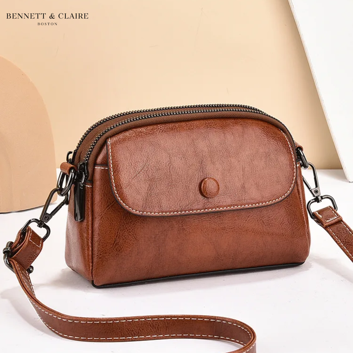 Crossbody-taske i cognacbrunt læderlook med klap, trykknap, kontrastsyninger og dobbelt lynlås, vist med den lange rem på lys baggrund. 1