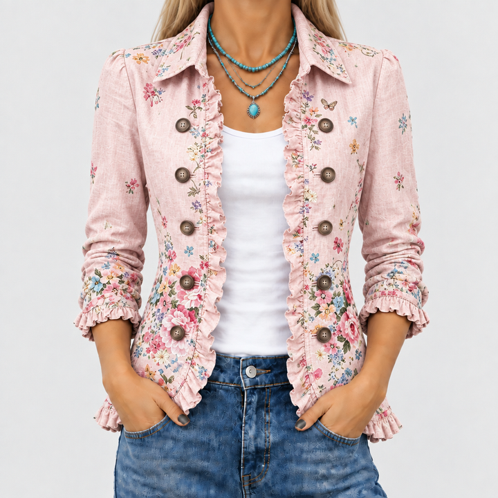 Nærbillede af den lyserøde blomstrede jakke med flæsekanter og knapper, båret åben over hvid top og jeans med turkis halskæde. 1