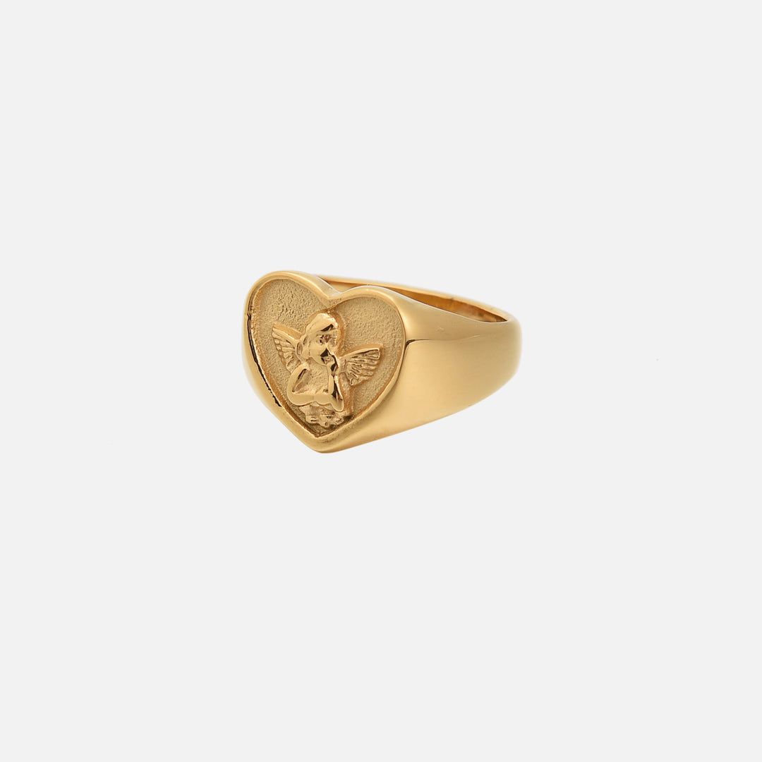 Guldfarvet ring set skråt fra siden mod lys baggrund, hjerteformet top med relief af en englefigur med vinger. 1