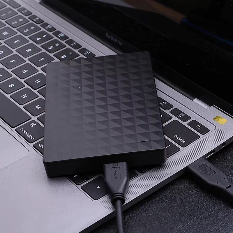 Sort harddisk med mønstret overflade, der ligger oven på en bærbar computers tastatur med tilsluttet USB-kabel 3