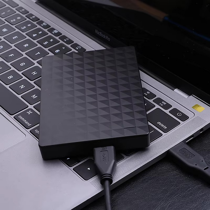 Sort harddisk med mønstret overflade, der ligger oven på en bærbar computers tastatur med tilsluttet USB-kabel 3