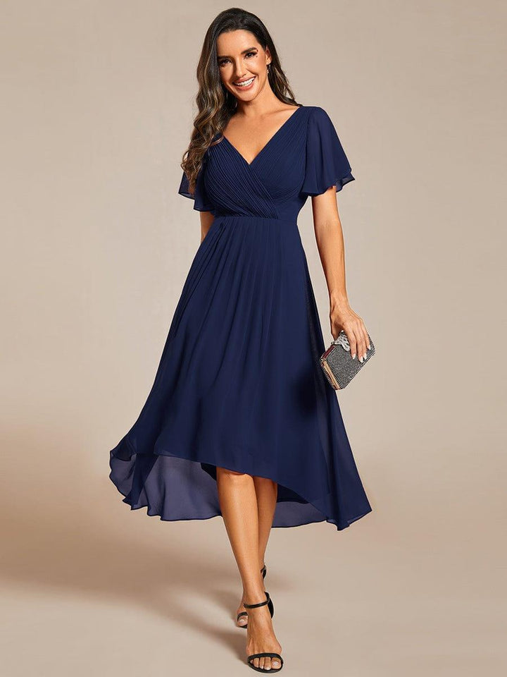 Model i marineblå chiffonkjole med plisseret V-hals og asymmetrisk kant set forfra, med glitrende clutch og sorte sandaler. 1