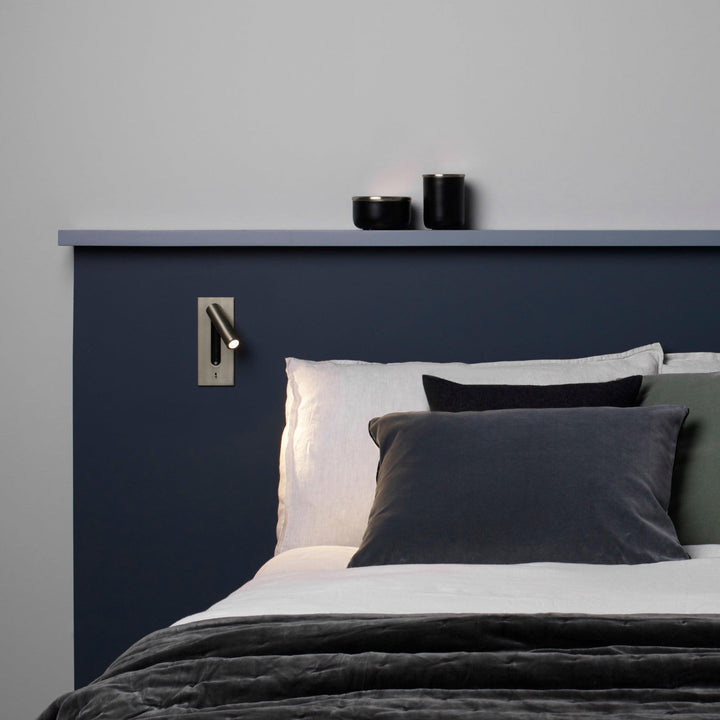 Væglampe i dæmpet metalfinish over en seng med blå sengegavl og grå puder 30