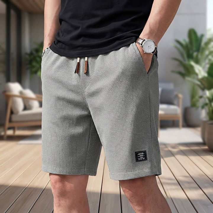 Mand i sort t-shirt og ur bærer grå vaffelstrikkede shorts med snøre i taljen og hænderne i lommerne på en terrasse. 6