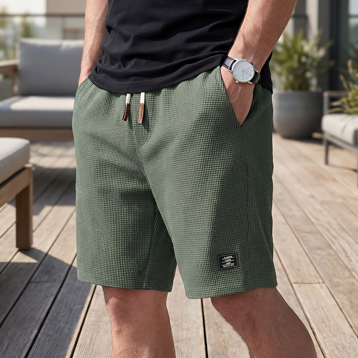 Mand i sort t-shirt og ur bærer grønne vaffelstrikkede shorts med hvidt snøre og lædertip, hånden i lommen, ude på en terrasse. 5