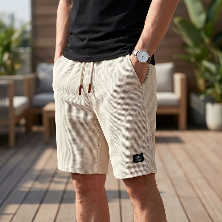 Mand i sort t-shirt og ur bærer cremefarvede vaffelstrikkede shorts med snøre og hænderne i lommerne foran en træterrasse. 8
