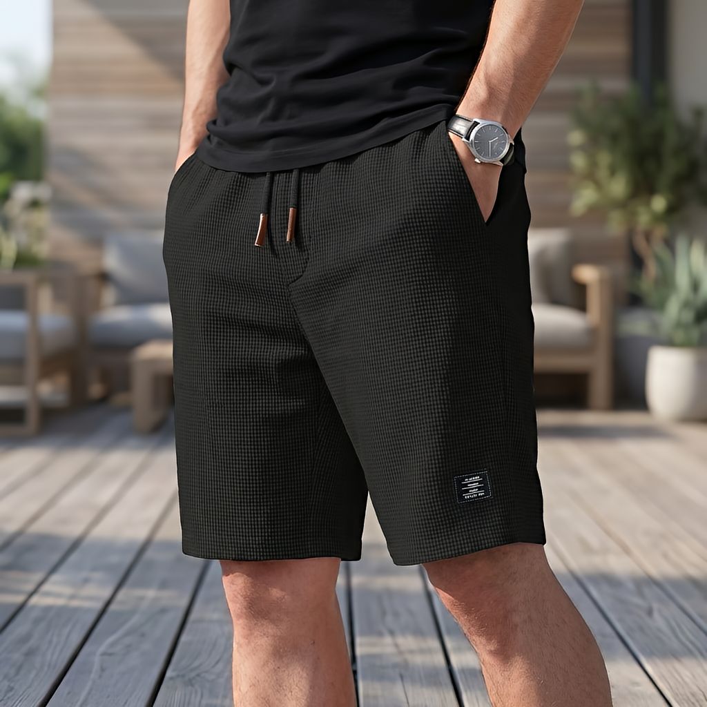 Mand i sort t-shirt og ur bærer sorte vaffelstrikkede shorts med snøre og lille mærke på benet på en trædæk-terrasse. 3