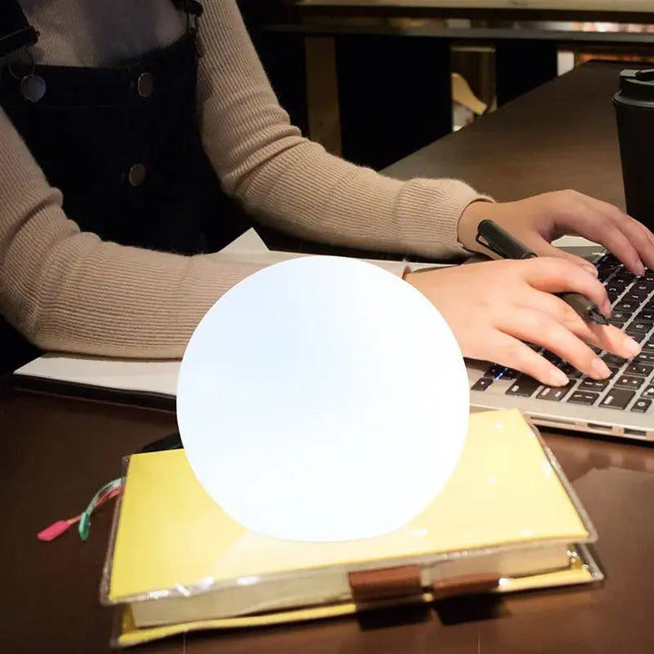 Hvid lysende kuglelampe på en notesbog på et skrivebord, mens en kvinde skriver på en bærbar computer. 7