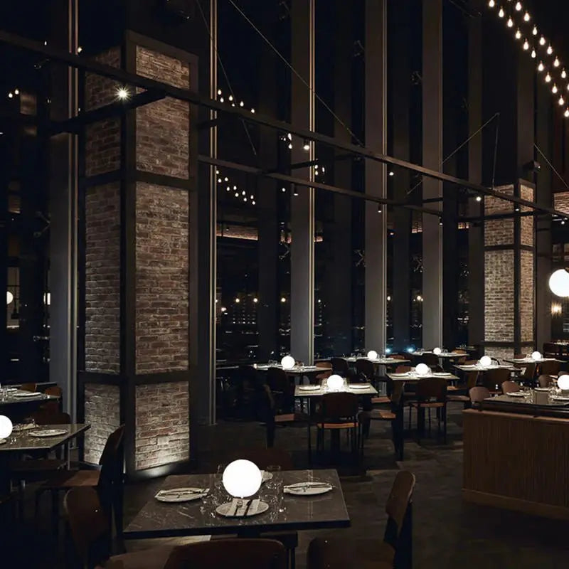 Flere kuglelamper lyser på bordene i en stor restaurant med murstenssøjler og mørk indretning. 8