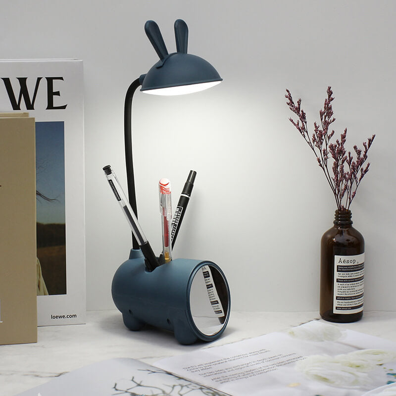 Tændt mørkeblå kaninlampe med tre penne i sokkelen og spejl foran, på et skrivebord med bøger og tørrede blomster. 9