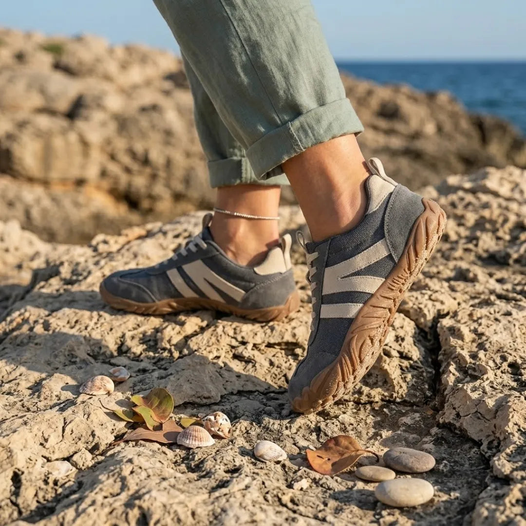 Grå sko med beige striber båret af model på klippekyst, med havet i baggrunden og små muslingeskaller foran. 2