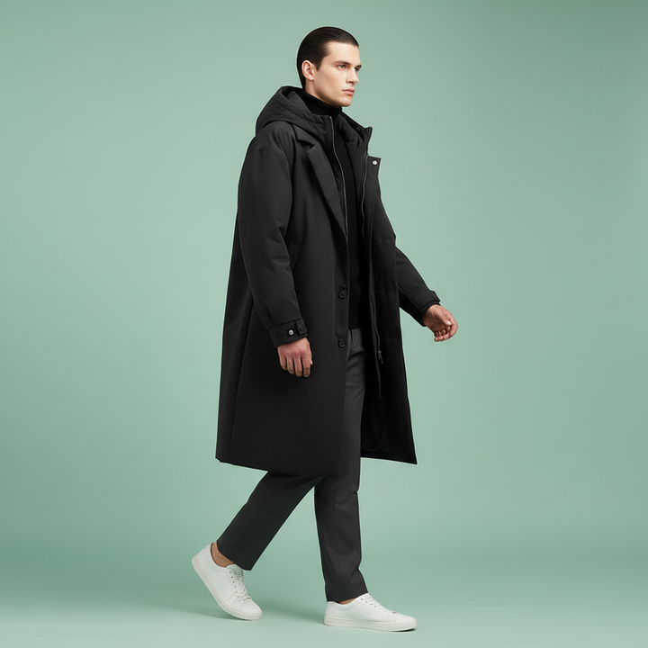 Model i åben sort frakke med hætte over sort trøje, grå bukser og hvide sneakers, gående mod mintgrøn baggrund. 7