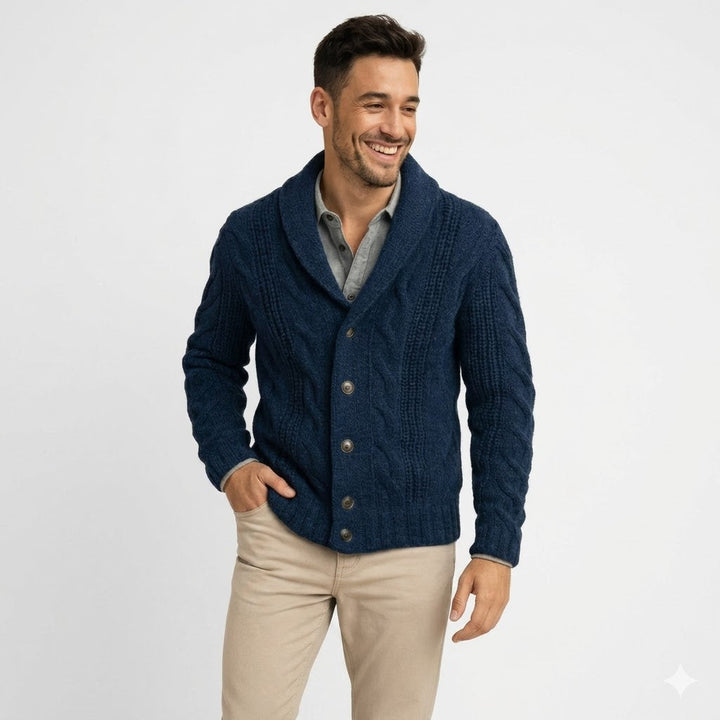 Smilende mand i marineblå flettestrikket cardigan med sjalskrave og knapper, med hånden i lommen på beige bukser, set fra livet og op. 1