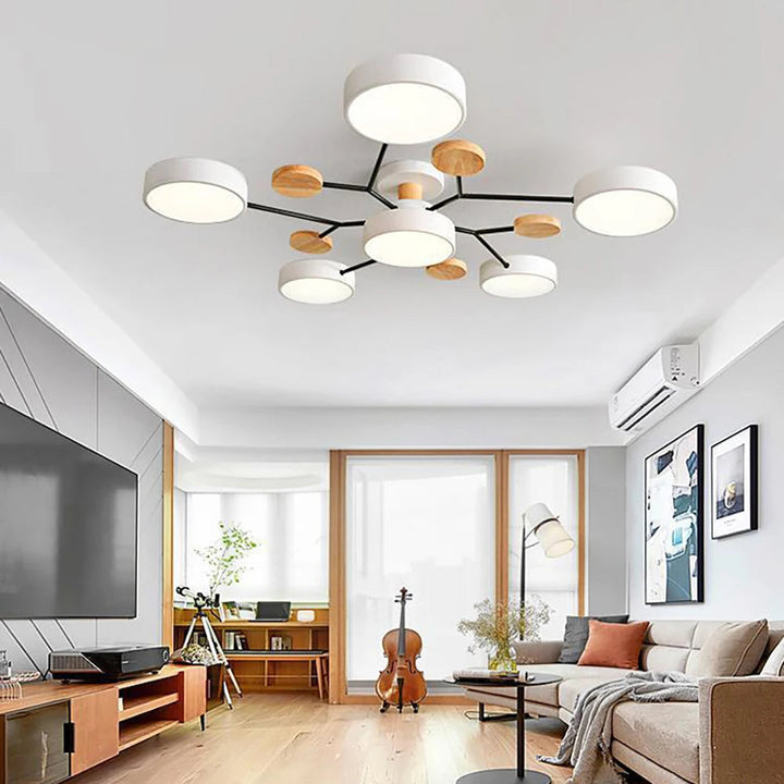 Hvid loftlampe med seks runde skærme, sorte arme og trædetaljer, set i en lys stue med tv-bord, cello og sofa. 3