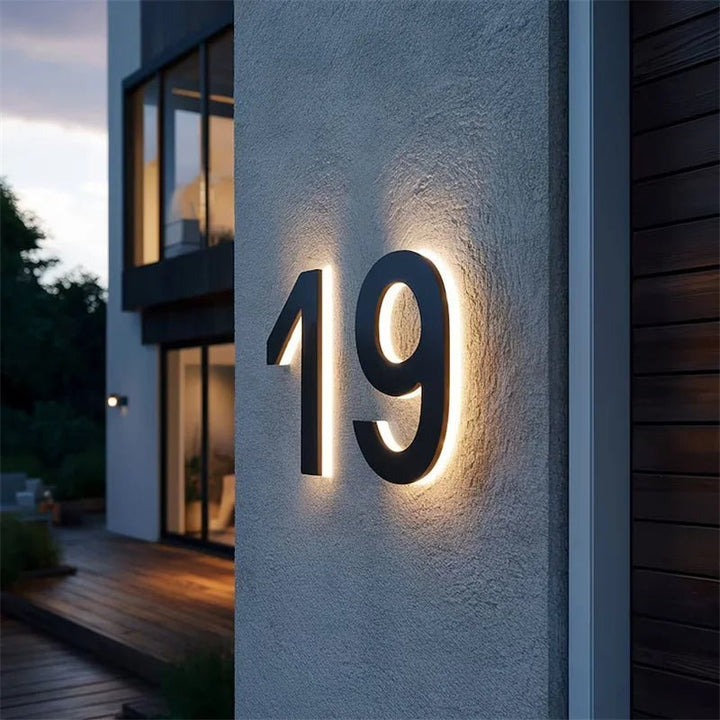 Sorte bagbelyste husnumre på en grå pudset væg i aftenlys, med glasfacade og træterrasse i baggrunden. 3