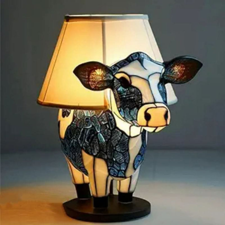 Lampe formet som en ko i blå og hvide mosaikfelter, med lys lampeskærm bag hovedet på rund sort sokkel. 12