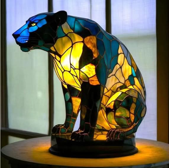 Siddende panter-lampe i blå, grønne og gule mosaikfelter, tændt på rund sokkel foran et vindue. 16