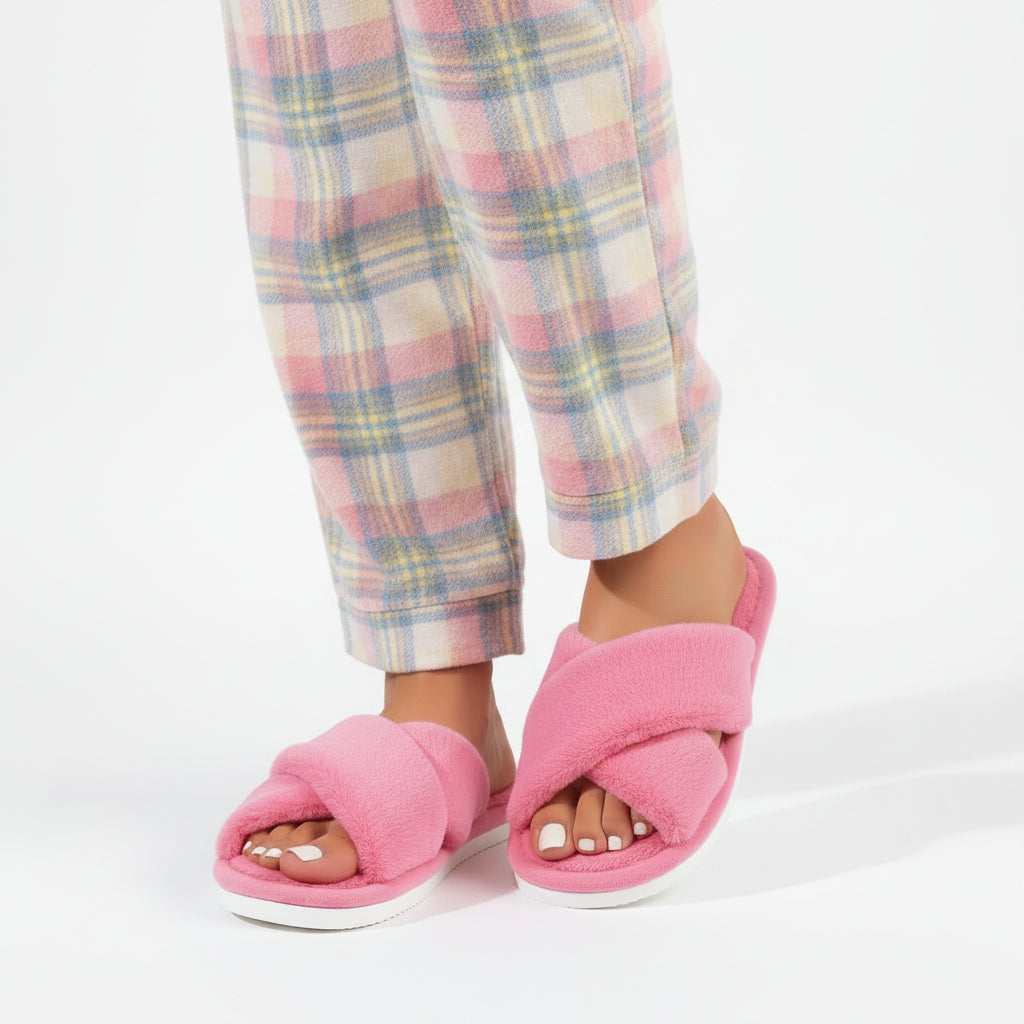 Lyserøde plyshjemmesko med krydsede remme båret af en model i ternede pyjamasbukser i lyse farver. 2