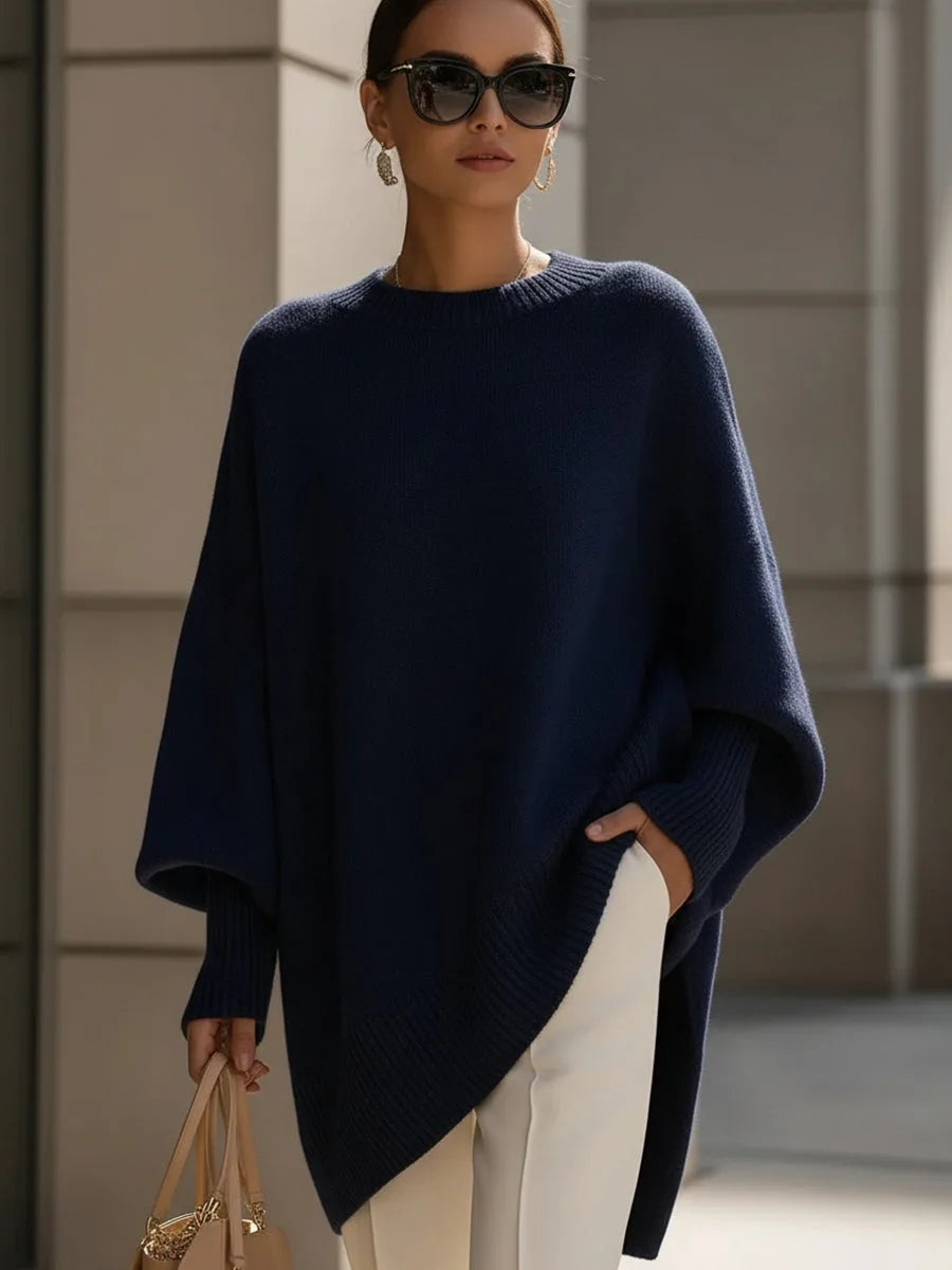Model i mørkeblå oversize strikbluse med asymmetrisk kant, solbriller, hvide bukser og taske. 8