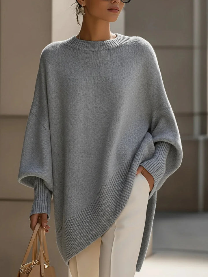 Model i grå oversize strikbluse med ribkant og hånden i lommen på hvide bukser. 9