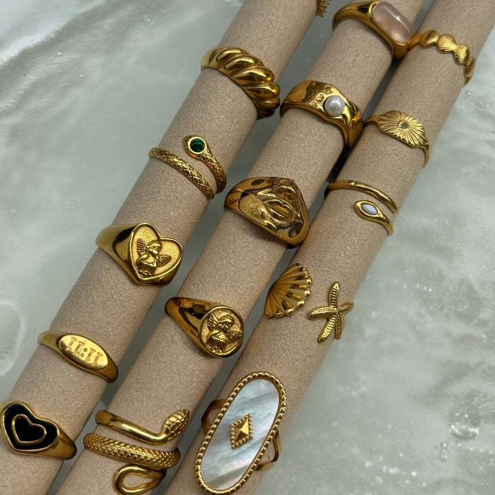 Flere guldfarvede ringe stillet på beige papruller, blandt dem hjerteringen med engel, sammen med slange-, perle- og skalringe. 5