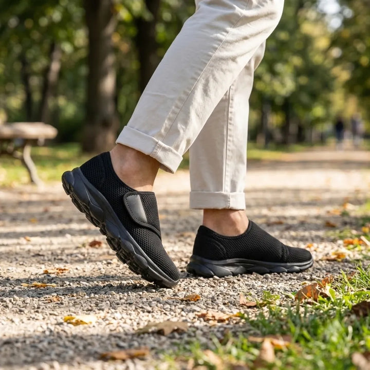 Sorte mesh-sko med velcrostrop og sort sål båret af en person i lyse bukser på en grusvej i en park. 11