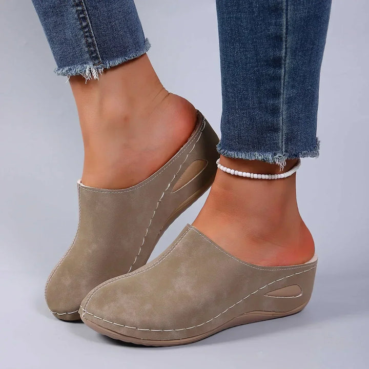Beige-grå tøfler med kileformet sål båret af en model med jeans og perlefodlænke, set skråt forfra. 3