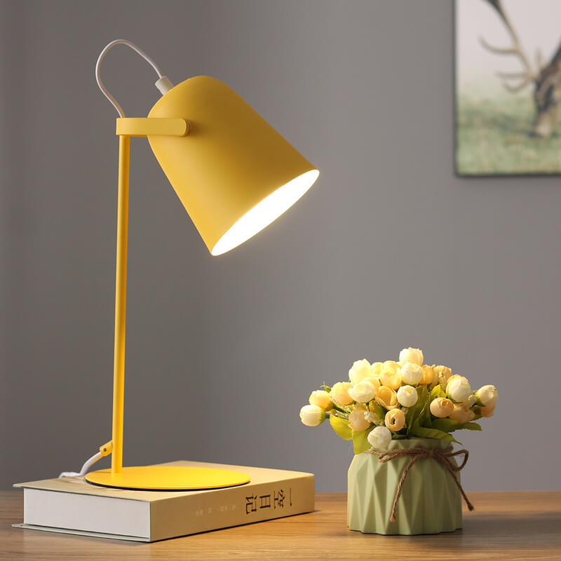 Tændt gul skrivebordslampe på en bog ved siden af en vase med blomster, med et indrammet motiv på væggen bagved. 7