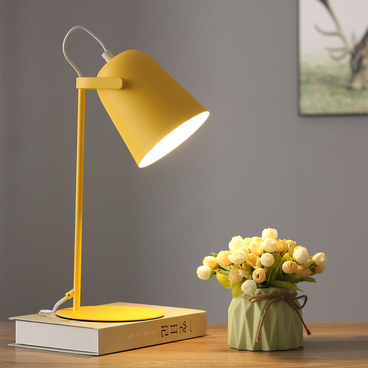 Tændt gul skrivebordslampe på en bog ved siden af en vase med blomster, med et indrammet motiv på væggen bagved. 7