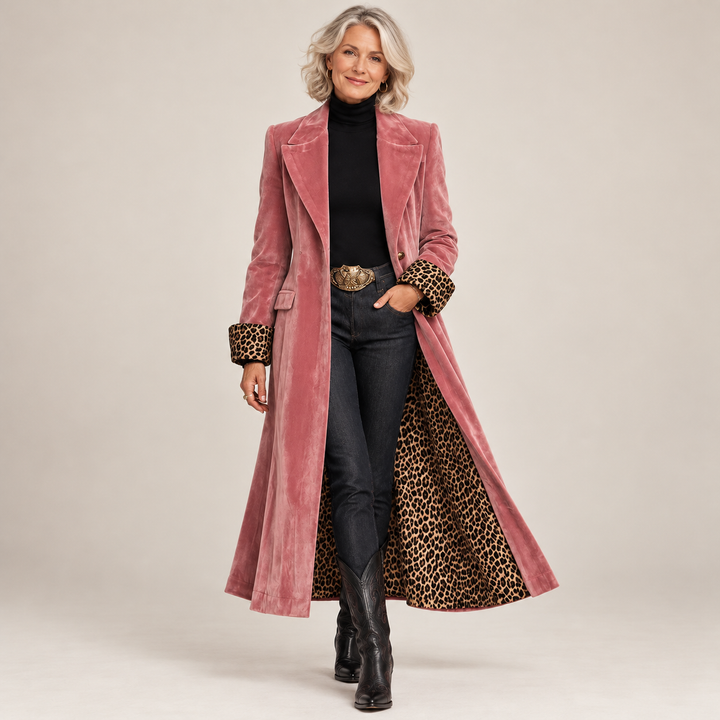 Smilende kvinde i hel figur i den lyserøde fløjlsfrakke åben over sort rullekrave, jeans og cowboystøvler, med leopardfor synligt. 1