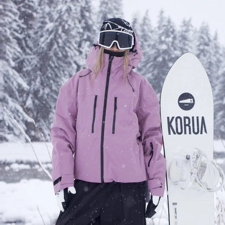 Model i lyslilla jakke med sorte lynlåse og lommer står i snefald og holder et hvidt snowboard ved siden af sig. 3