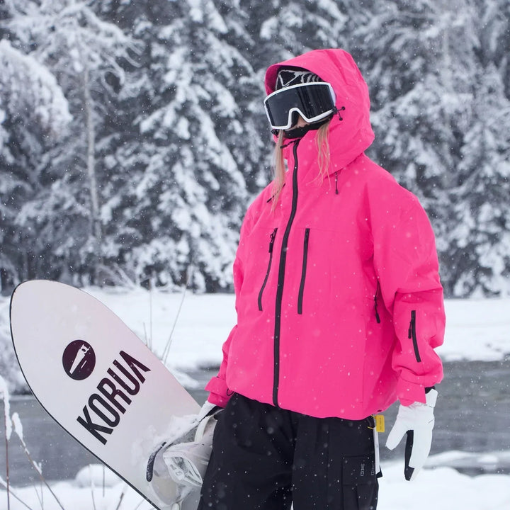 Model i skarp pink jakke med hætte, skibriller og sorte bukser går med et snowboard under armen i snedækket skov. 5