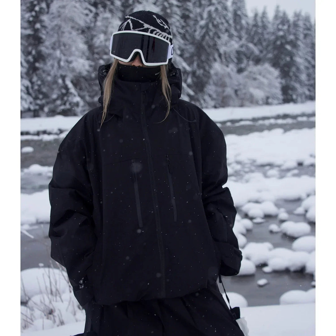Model i sort jakke med høj krave og hætte, skibriller og hue står ved en delvist frossen flod i sneen. 2