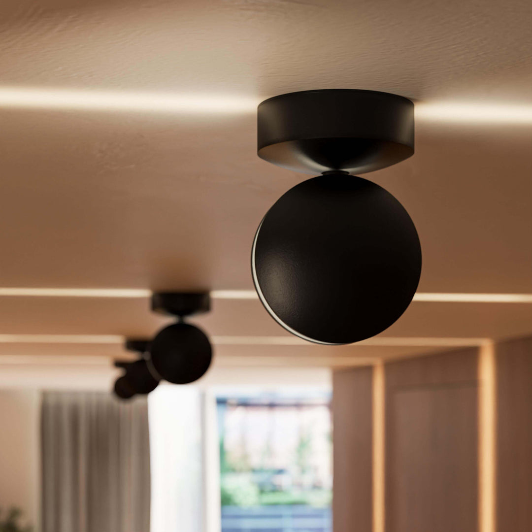 Nærbillede af en sort loftlampe med rundt hoved, der kaster en vandret lysstribe langs det varme loft; en anden lampe sløret bagved. 11