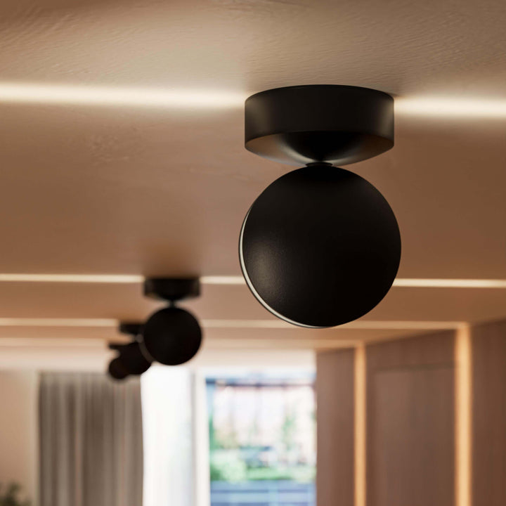 Nærbillede af en sort loftlampe med rundt hoved, der kaster en vandret lysstribe langs det varme loft; en anden lampe sløret bagved. 11