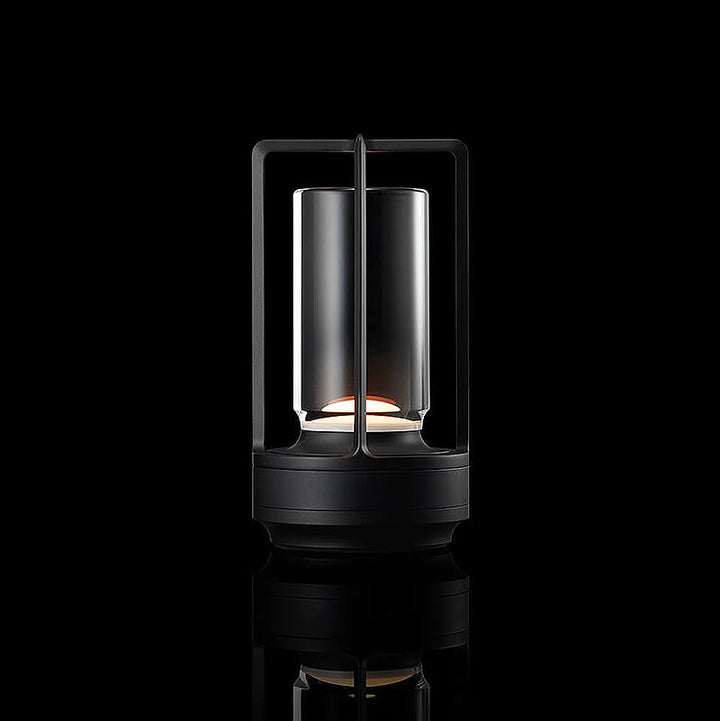 Lampe med sort metalstel og klar glasskærm, tændt mod sort baggrund med spejling. 11