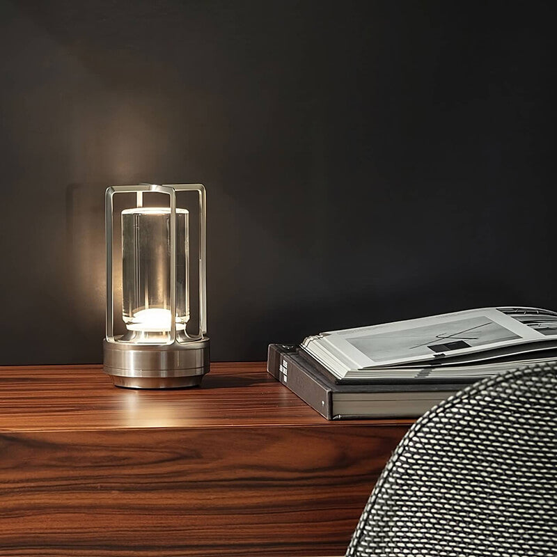 Sølvfarvet lampe med tændt lys på et træskrivebord foran en mørk væg, ved siden af opslåede bøger. 5