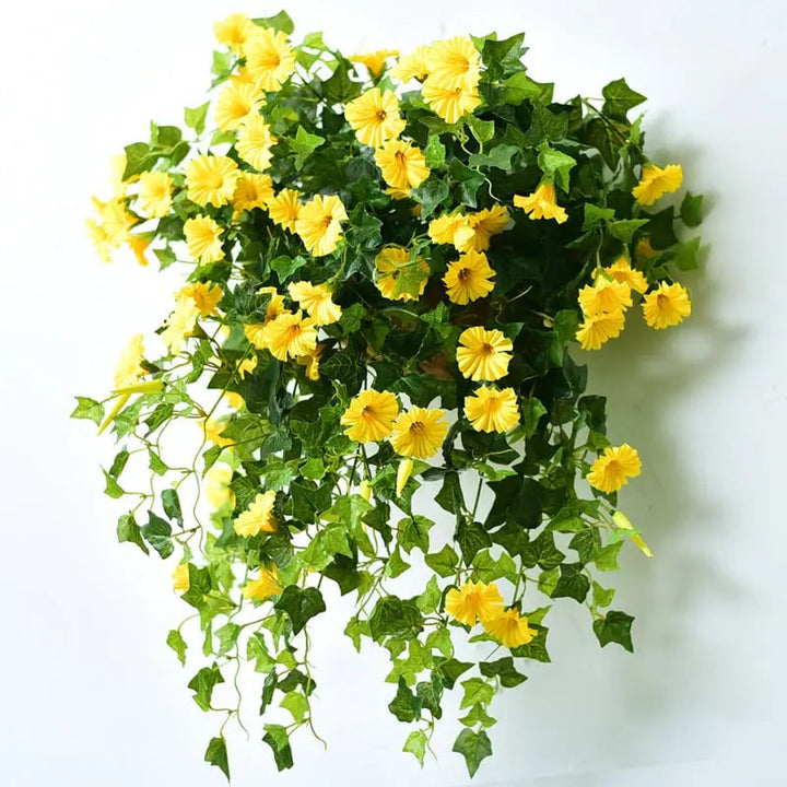 Gule kunstige blomster med grønne blade og lange nedhængende ranker, vist på en hvid væg. 5