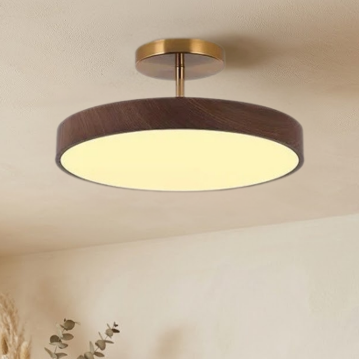 Tændt lampe med mørkt trælook og guldfarvet stel set nedefra og lidt fra siden, med varmt lys og dekorationsgrene i hjørnet. 3
